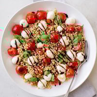 Freshly cooked pearled couscous tossed with halved cherry tomatoes, mozzarella balls, and torn basil leaves on a white platter, drizzled with balsamic glaze.  
